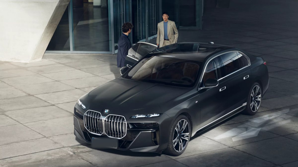 A man holding the rear door of a black BMW 7 Series open as a passenger walks out of a building towards the car